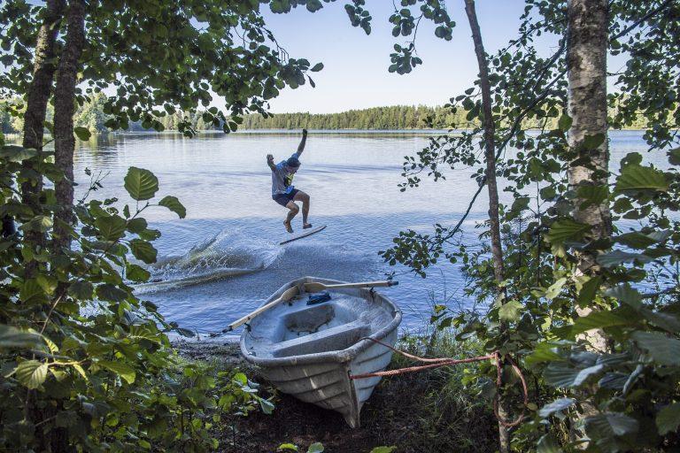 Lehtipuiden välistä, soutuveneen yli näkyy järvelle. Nuori mies hyppää juuri skeittilaudan tapaisella laudalla aallokossa., kädet ylhäällä.