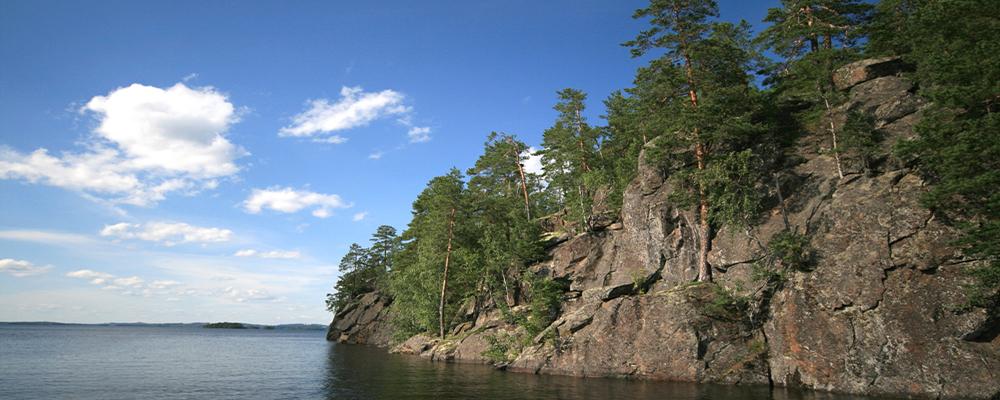 kesäinen maisemakuva, jossa näkyy järvestä nouseva jylhä kallioseinämä. Seinämällä kasvaa vihreitä puita. Sää on pilvipoutainen.
