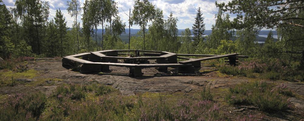 Pyöreähköön muotoon rakennettu tulipaikka, jota ympäröi kesäinen harjumaisema.