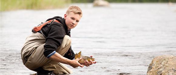 Nuori, noin 14-vuotias vaaleatukkainen poika on kyykyssä matalassa jokivedessä pukeutuneena kahluuhousuihin ja pitää käsissään lohikalaa. 