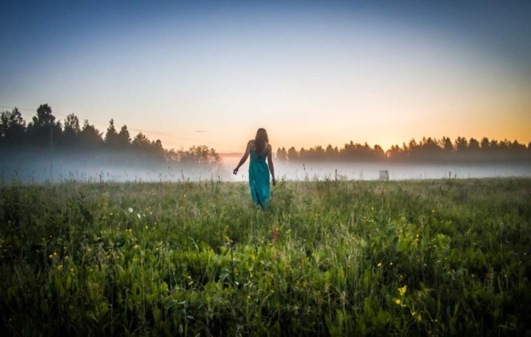 Nainen kävelee kesäisellä pellolla, jolla leijuu usvaa.